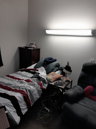 A cozy but cluttered bedroom with a neatly made bed, a chair, and various items scattered around.