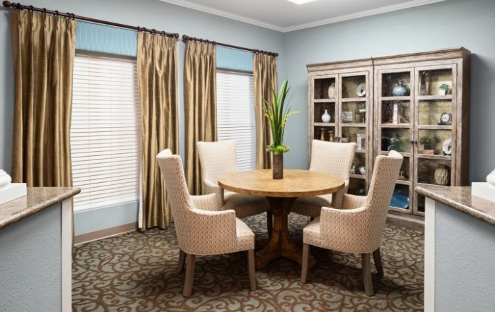 A stylish dining room with a round wooden table surrounded by four upholstered chairs. The room features elegant curtains, a decorative cabinet, and a potted plant centerpiece.