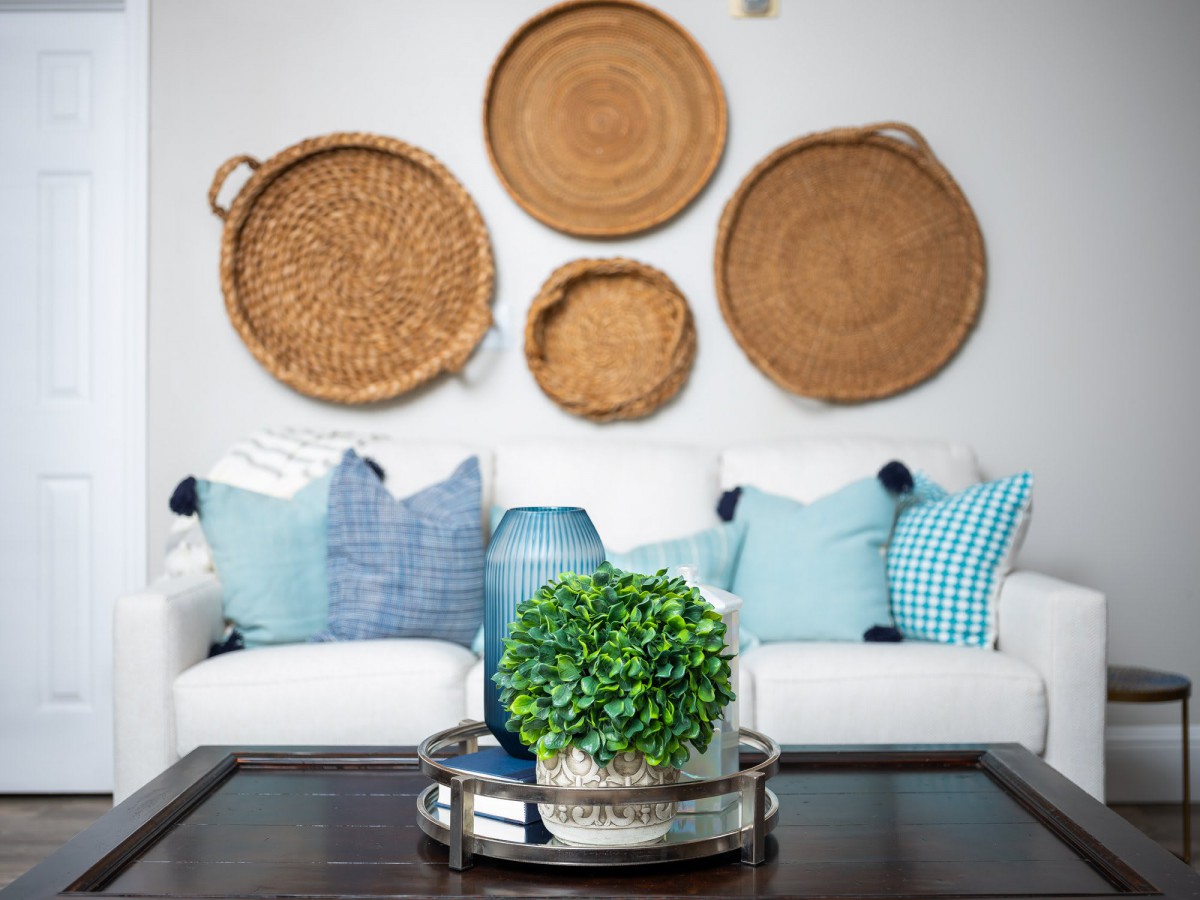 A cozy living room featuring a white couch with blue pillows, adorned with wicker baskets as wall art and a coffee table centerpiece with green foliage.