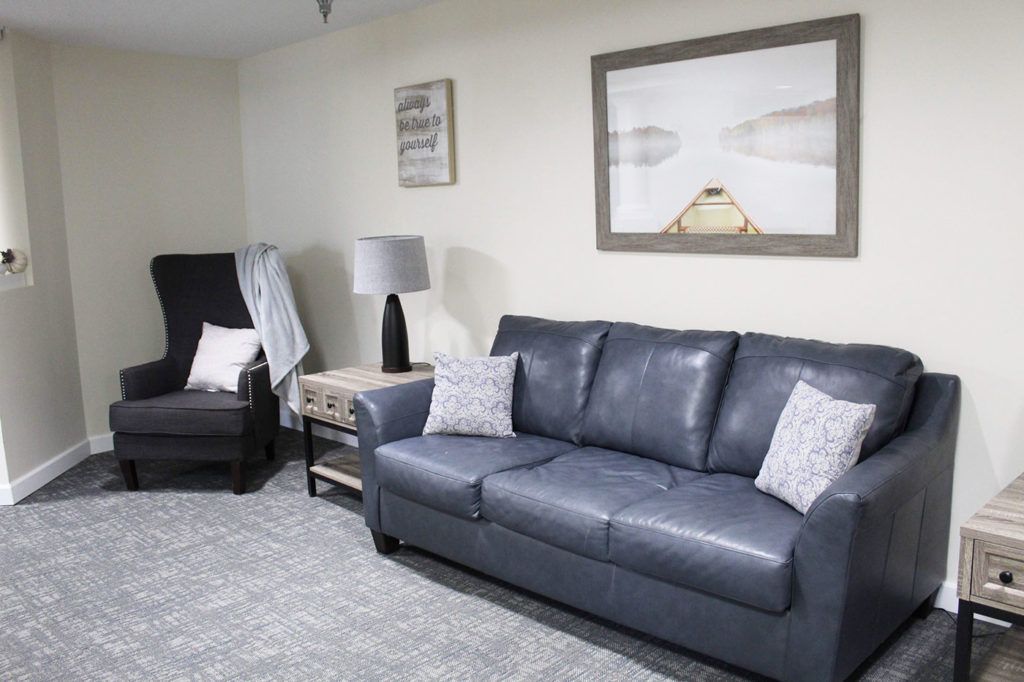 A cozy living room with a blue sofa, an armchair, artwork on the wall, and a lamp on a side table.