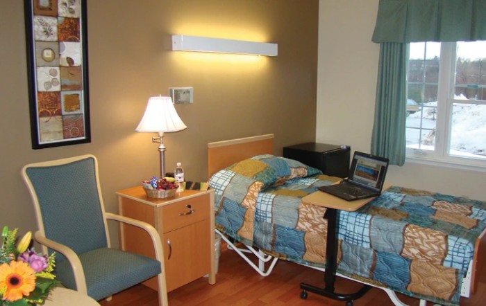 A hospital room featuring a bed, side table with a lamp, chair, and a laptop on a small table.