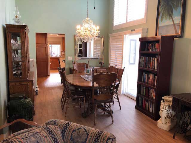 A cozy dining room with a wooden table and chandelier, alongside a bookshelf and large windows.
