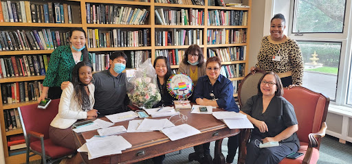 A group of individuals celebrating with flowers and a decorative plate in an office setting surrounded by books.