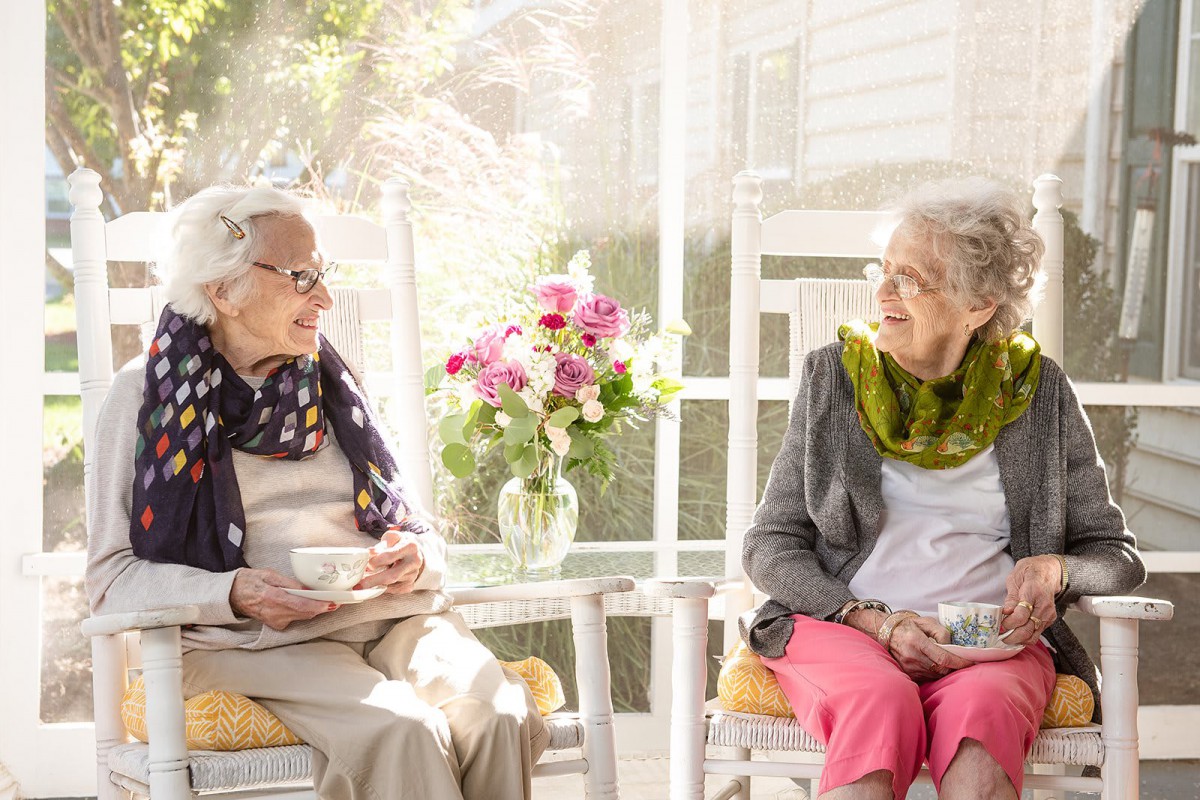 Two elderly women enjoying a conversation in a sunlit room with flowers on the table.