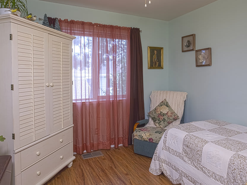 Cozy bedroom with classic armoire, bed, and a comfortable chair next to a window with sheer curtains.