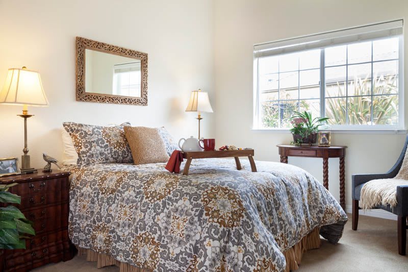 A cozy bedroom with a patterned bedspread, bedside lamp, and a window letting in natural light.