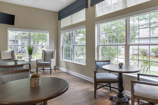 A cozy living room with large windows, allowing natural light to brighten the space, featuring comfortable chairs and round tables.