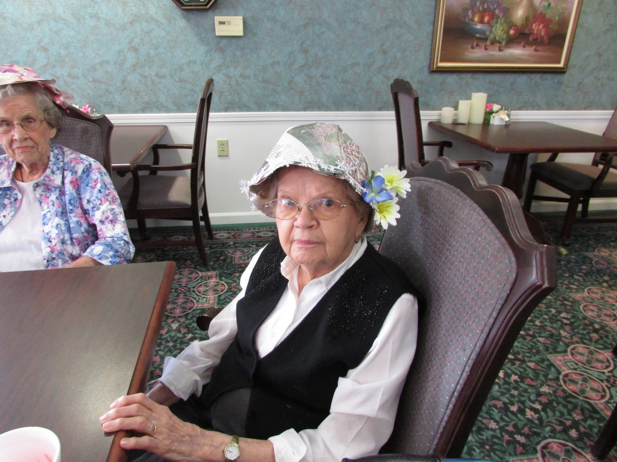An elderly woman wearing a decorative hat sits at a table in a dining area.