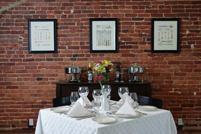 A neatly set dining table with wine glasses and white napkins against a brick wall adorned with framed art.