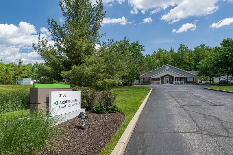 An office building with a landscaped entrance and a visible sign along the driveway on a clear day.