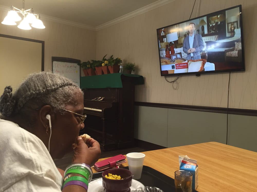 An elderly person enjoys a snack while watching television at a dining table.