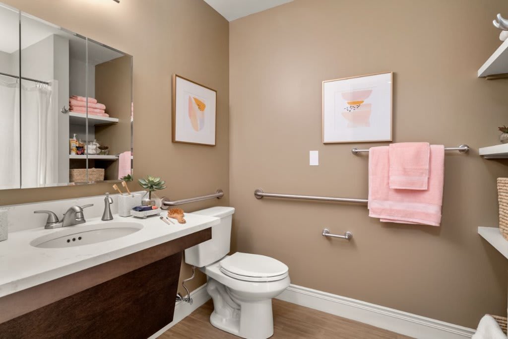A modern bathroom with neutral walls, featuring a toilet, sink, mirror, and decorative wall art. Pink towels add a soft touch.