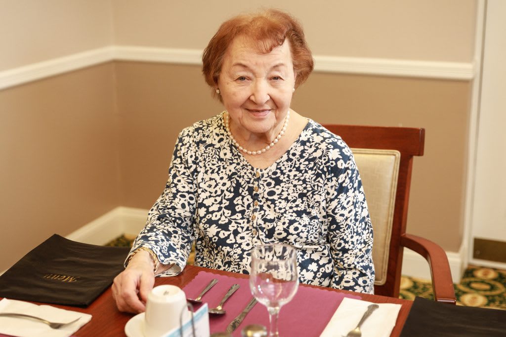 An elderly woman sitting at a dining table in a restaurant, smiling at the camera.