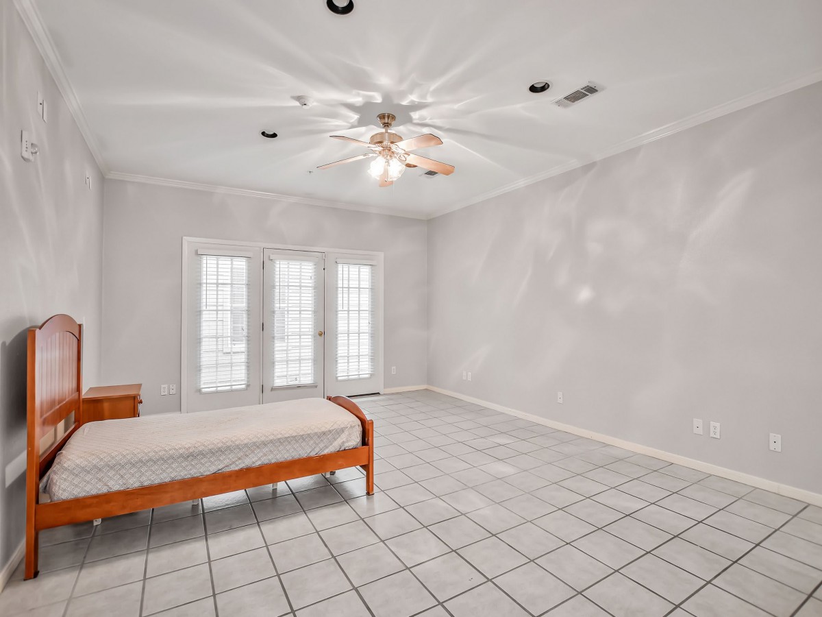 A spacious bedroom with a single bed, white walls, and tiled flooring, featuring large windows and a ceiling fan.