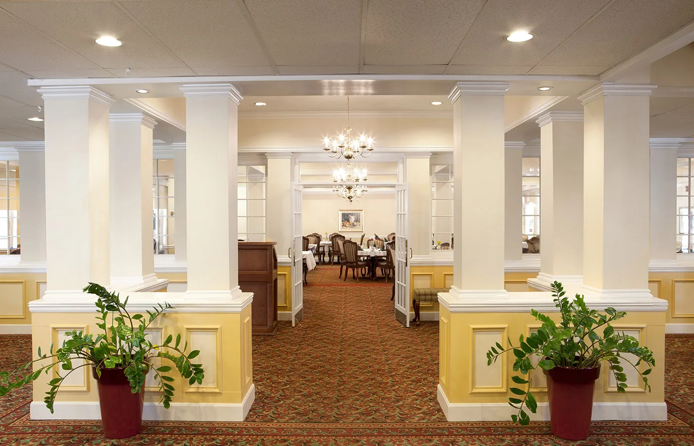 An elegant dining room with chandeliers and neatly arranged tables, featuring a carpeted floor and decorative plants.