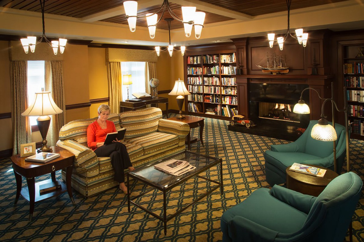 A cozy library room with a person reading on a couch, surrounded by bookshelves and a fireplace.