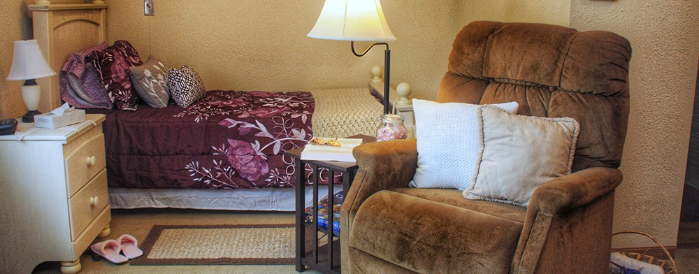 A cozy living room featuring a brown sofa with white pillows, a lamp, and a side table next to a bed with a patterned spread.