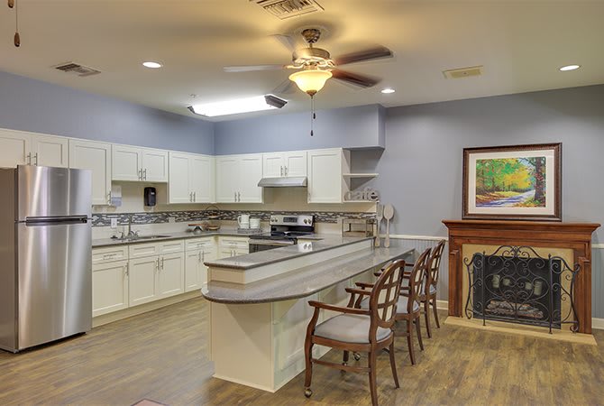 A spacious kitchen with white cabinets, a large countertop equipped with bar stools, and a ceiling fan. A fireplace with framed artwork adds to the cozy ambiance.
