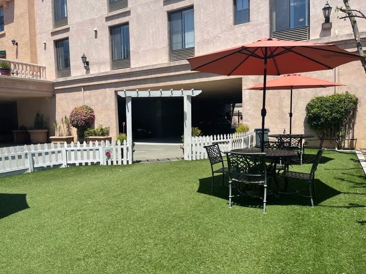 A sunny outdoor patio area with a table and chairs under an umbrella, surrounded by a green lawn and a white fence.
