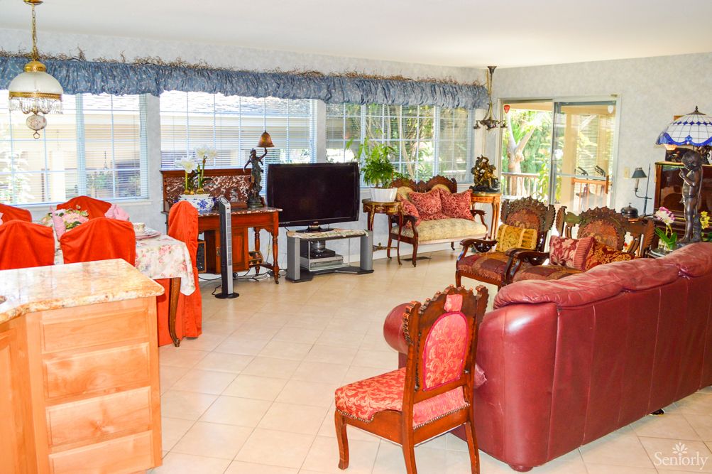 A spacious living room with various furniture pieces, including sofas, chairs, and a television, bathed in natural light from large windows.