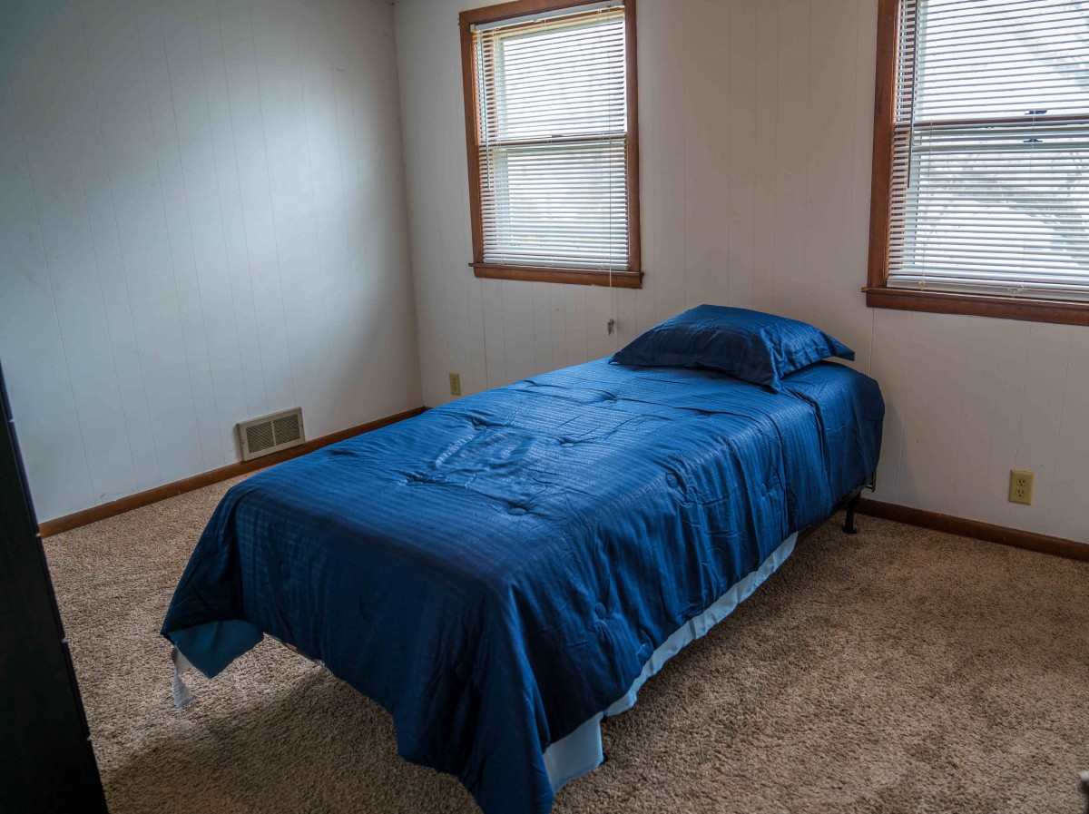 A minimalist bedroom with a single bed covered in blue bedding and two windows providing natural light.