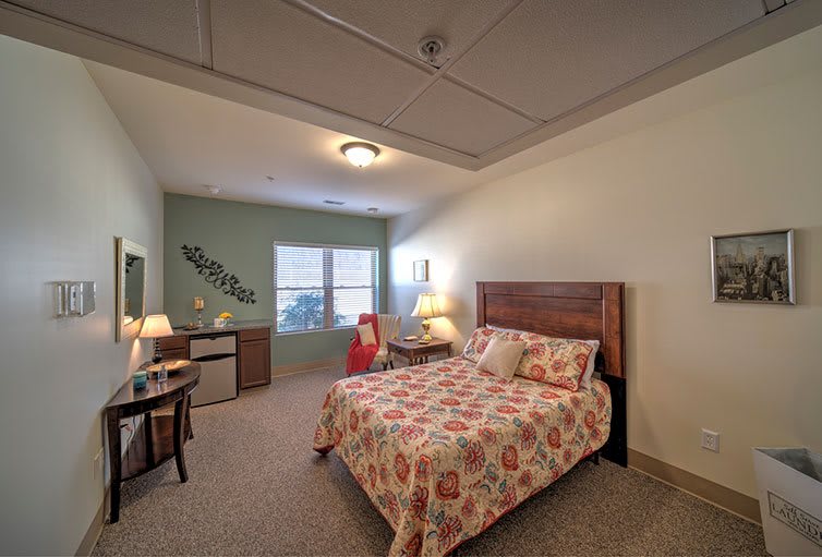 A cozy bedroom interior with a floral-patterned bedspread, a wooden headboard, ambient lighting from lamps, and a window providing natural light.