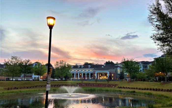 A serene evening view of a pond with a fountain and a nearby streetlamp, set against the backdrop of a sunset sky and distant buildings.