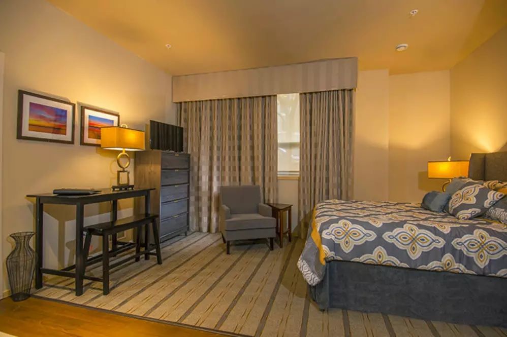A cozy hotel room featuring a bed, armchair, desk with a lamp, and patterned curtains.