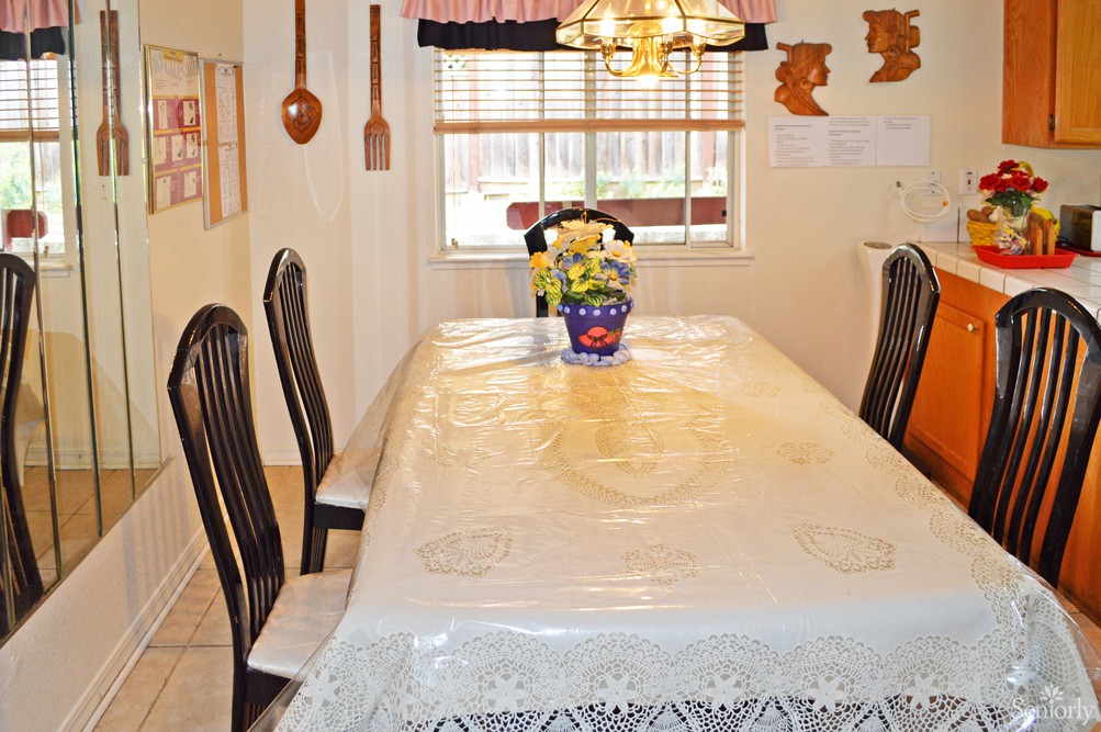 A dining room with a table covered in a lace tablecloth, surrounded by chairs and featuring a floral centerpiece under a chandelier.