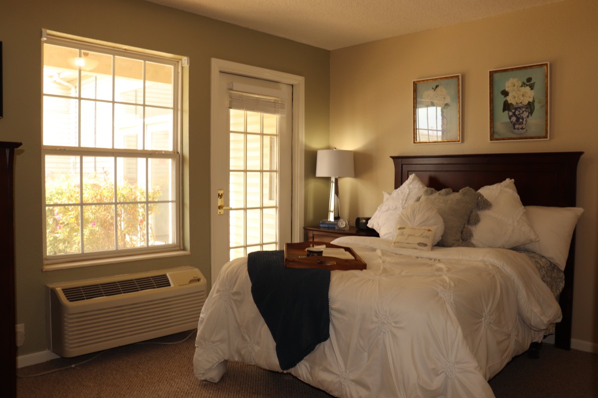 A cozy bedroom interior with a neatly made bed, soft lighting, and abundant natural light streaming through a large window.