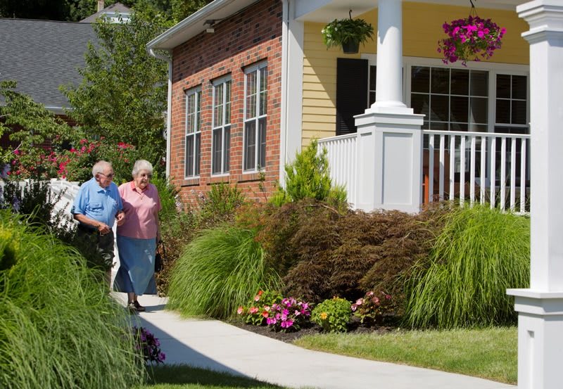 An elderly couple walks along a pathway beside a beautifully landscaped home with a welcoming front porch.
