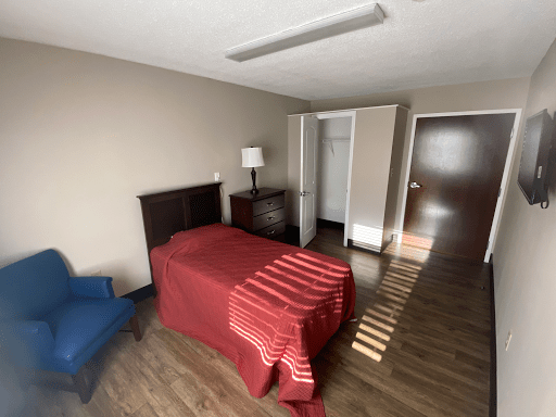 A minimalist bedroom featuring a single bed with a red blanket, a lamp on a bedside table, an open closet, and a wooden floor.