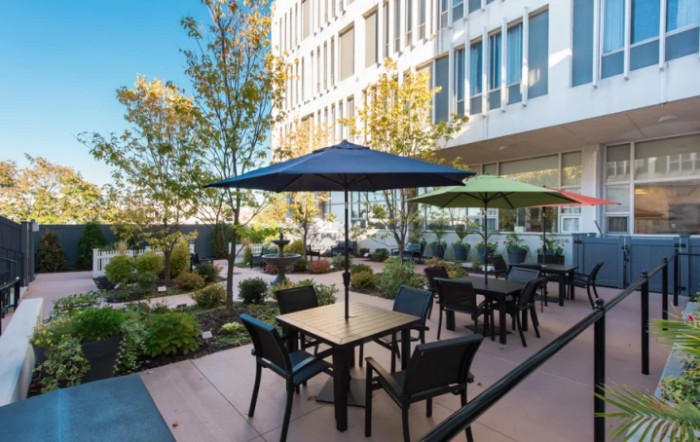 An outdoor patio area with tables, chairs, and umbrellas surrounded by trees and adjacent to a modern building.