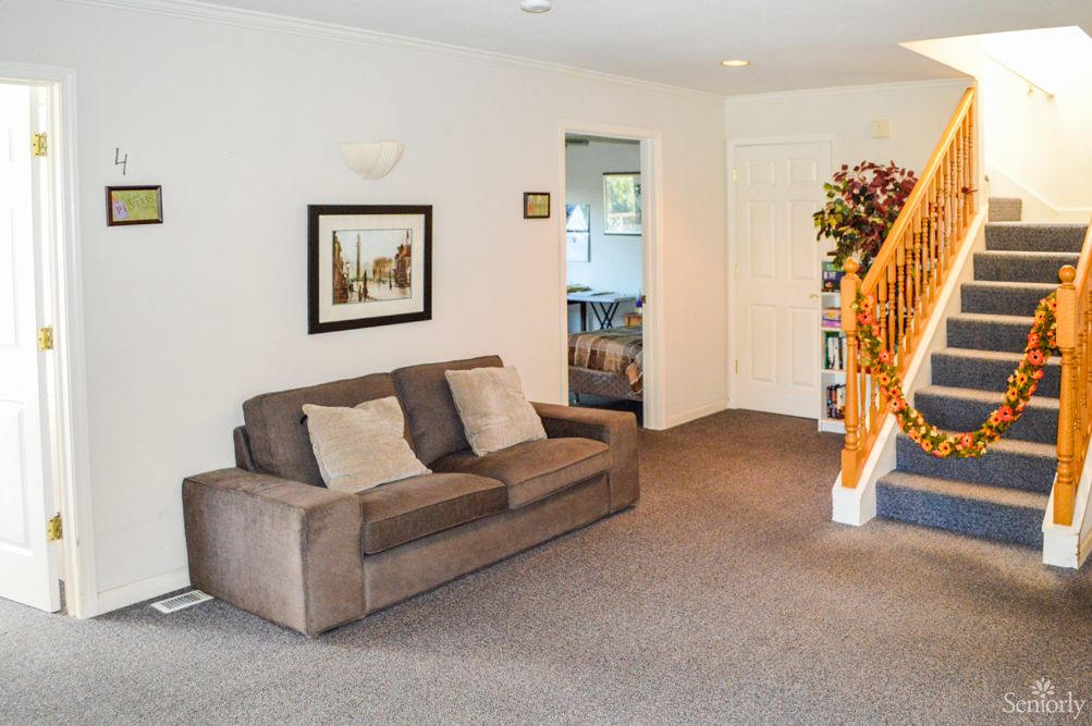 A cozy living room featuring a brown sofa, carpeted floor, artwork on the wall, and wooden stairs leading upstairs.