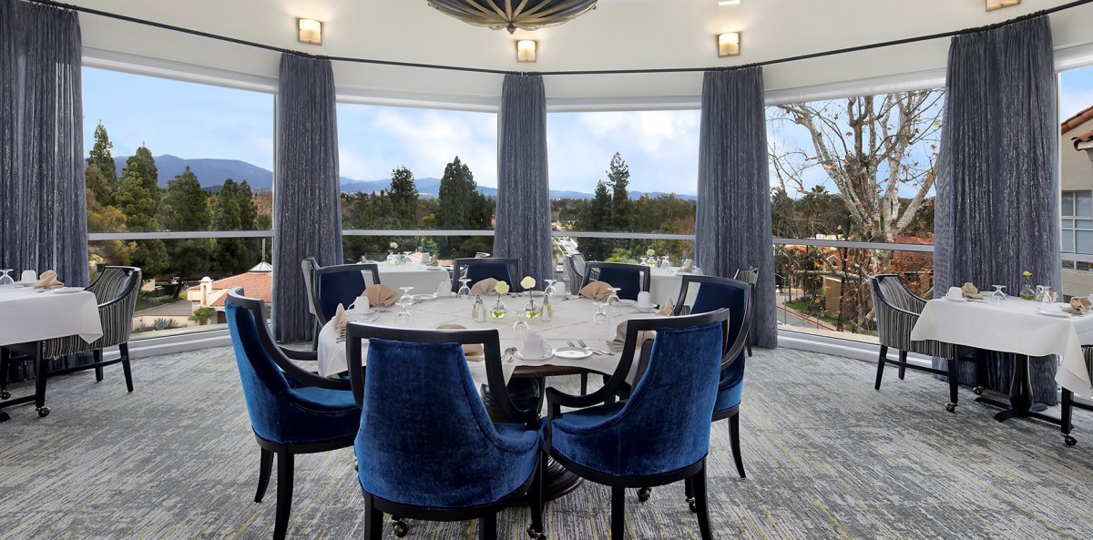 An elegantly decorated dining room featuring a round table surrounded by blue chairs, with large windows offering an outdoor view.