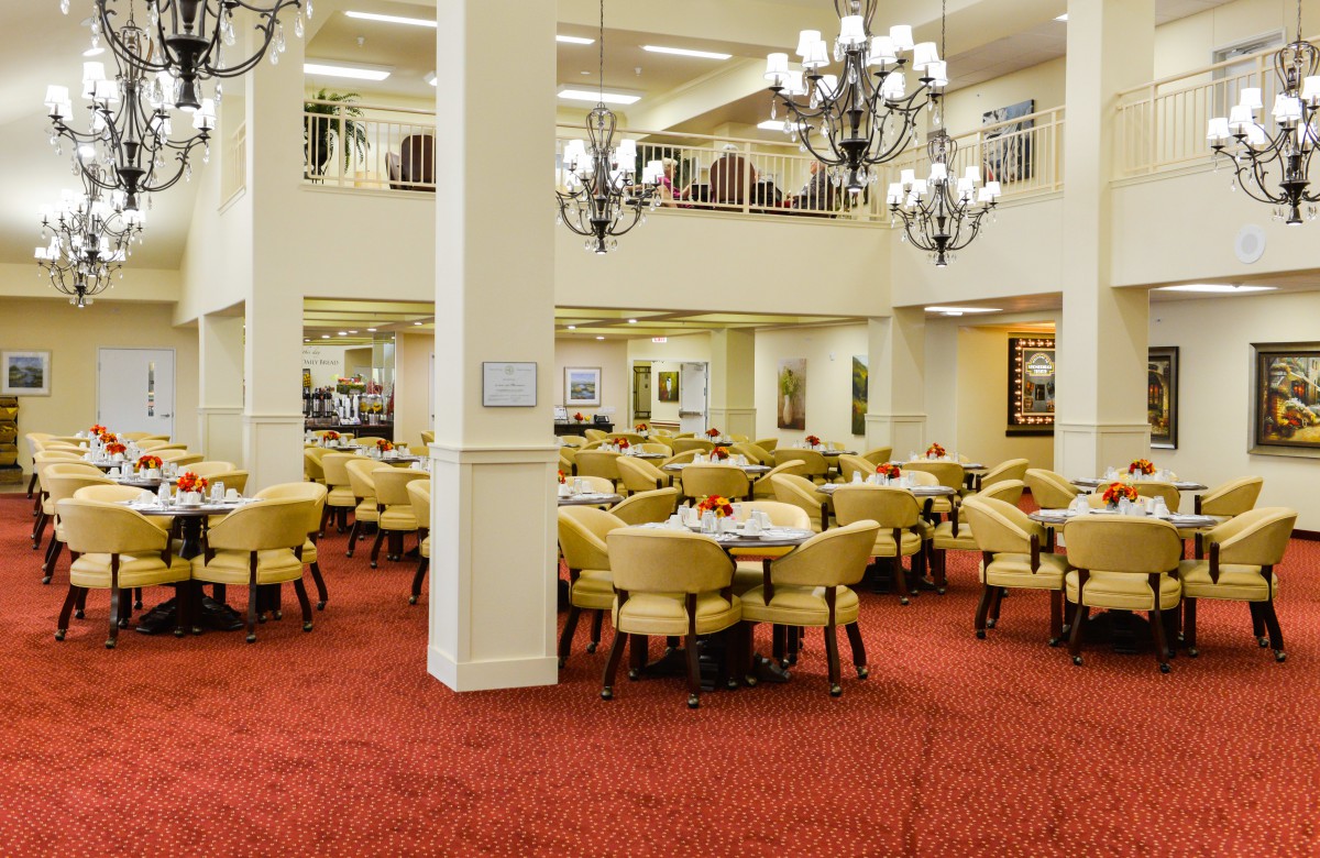A spacious dining room with elegantly arranged tables and chairs, featuring chandeliers and a red carpeted floor.