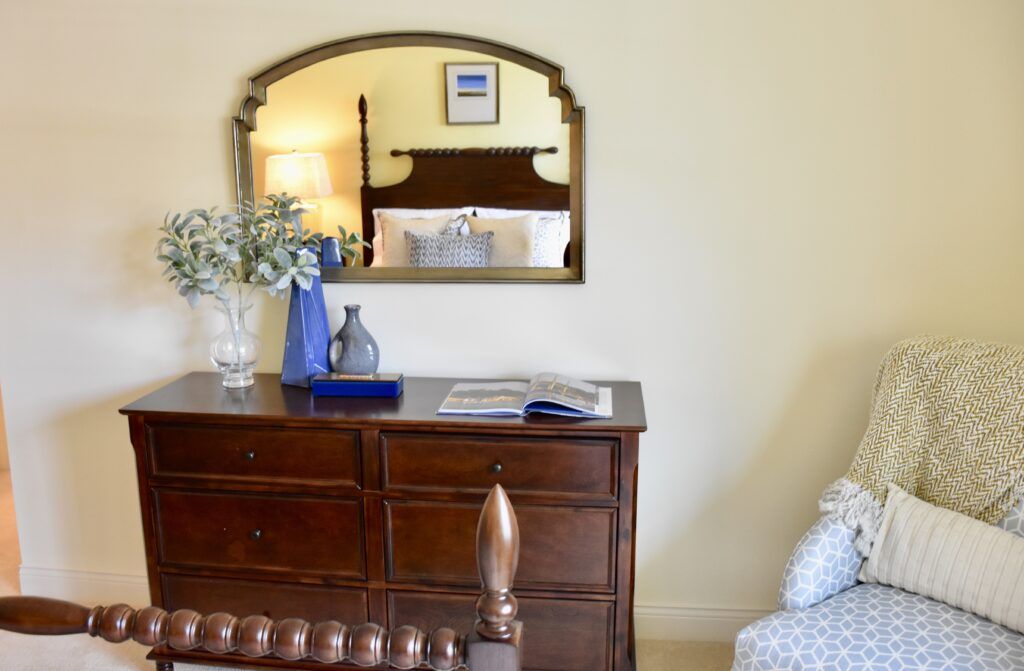 A cozy bedroom scene featuring a wooden dresser with decorative items, a mirror, and a glimpse of the bed.