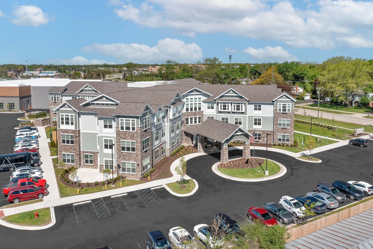 Aerial view of a modern residential building with a surrounding parking lot and landscaped areas under a partly cloudy sky.