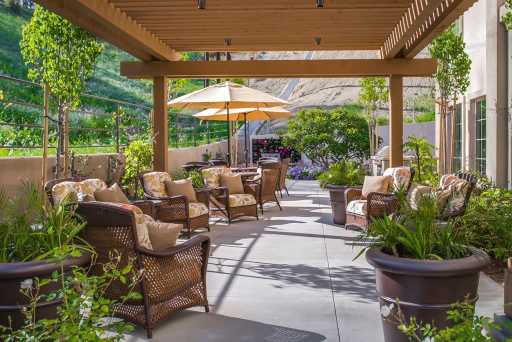 A cozy patio with comfortable seating under a pergola, surrounded by greenery and shaded by an umbrella.