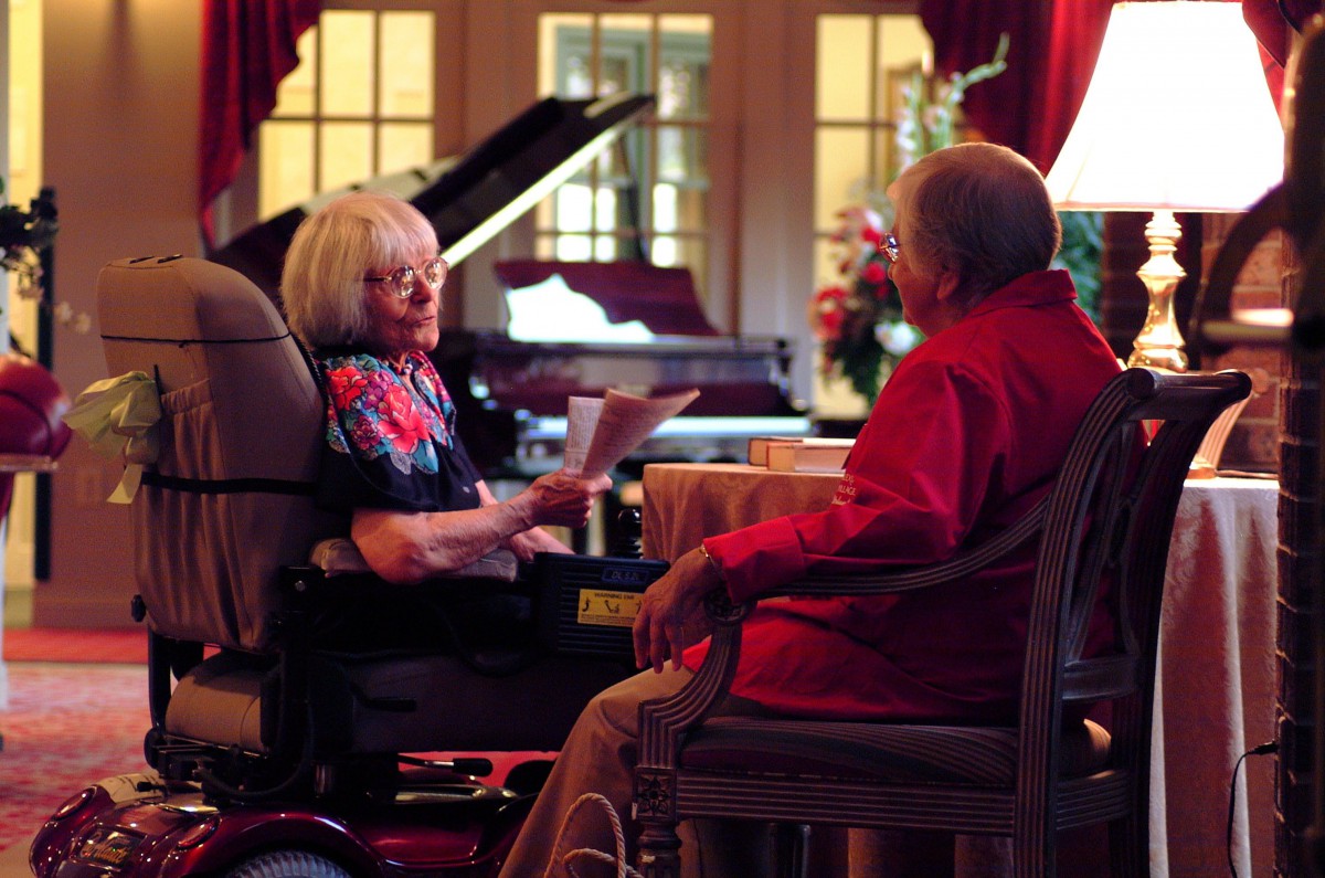 Two seniors engaging in a conversation in a cozy living room setting with a piano in the background.