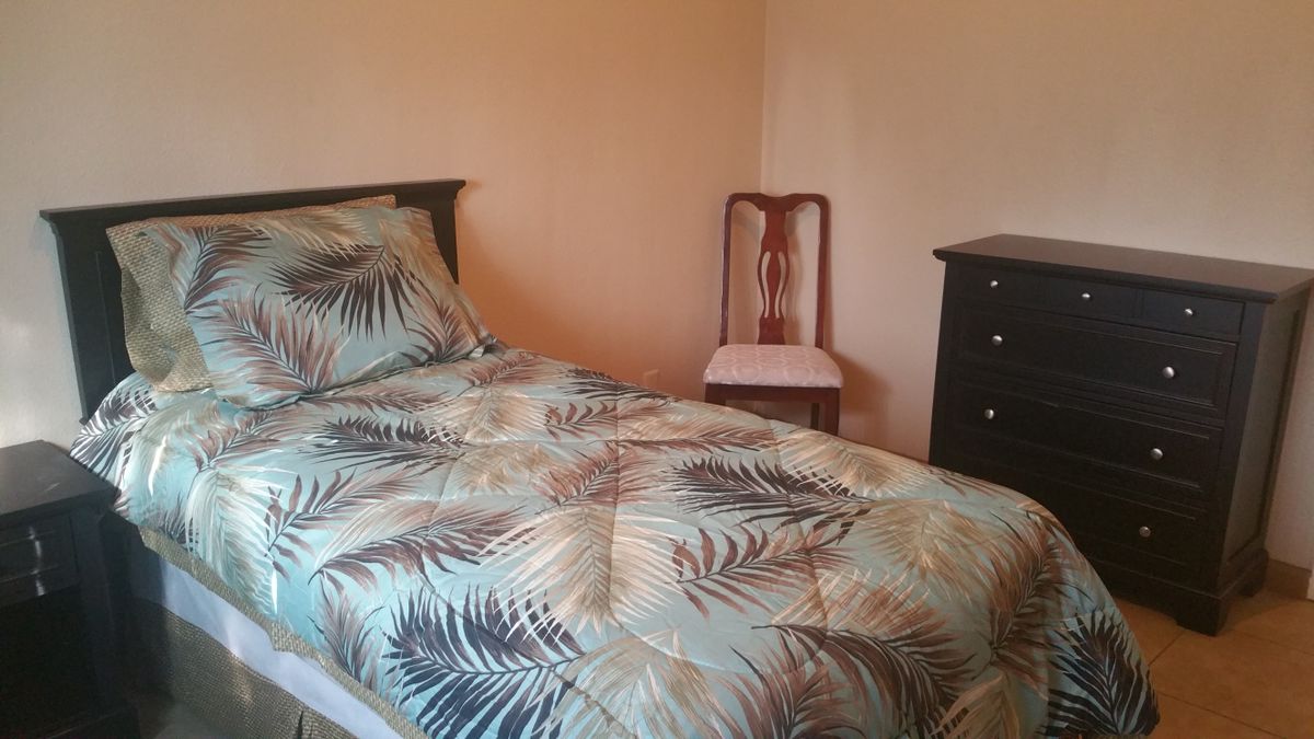 A cozy bedroom with a tropical-themed bedspread, a wooden chair, and a dark dresser against peach-colored walls.