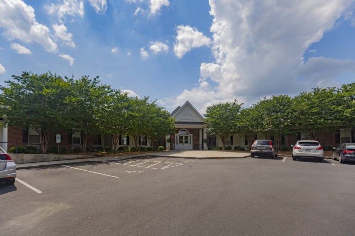 A building with a parking lot in front, flanked by trees and under a cloudy sky.