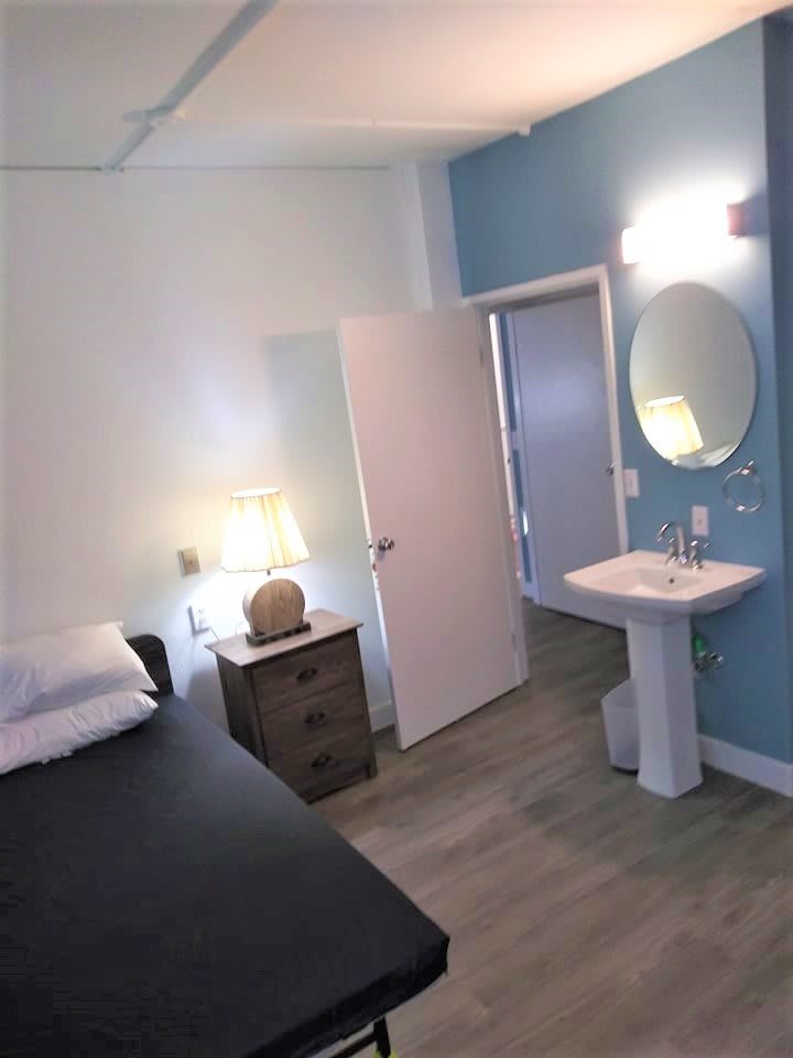 A minimalist bedroom with a bed, nightstand, lamp, and adjacent sink area.
