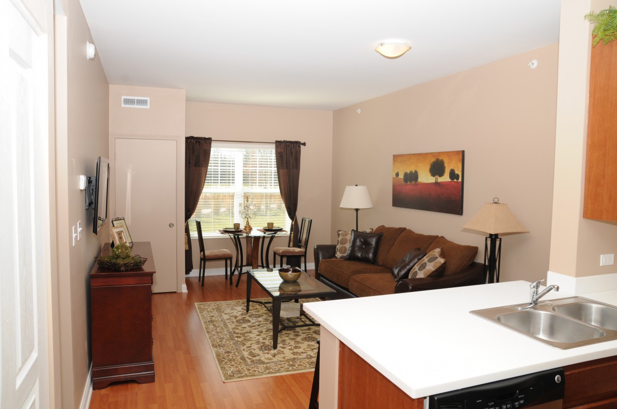 A cozy apartment living room with a sofa and dining area by the window, featuring warm tones and wood flooring.