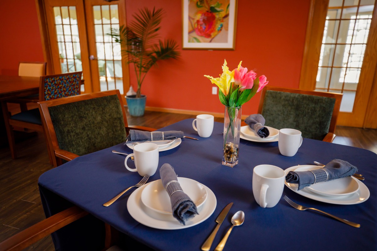 A dining table set for four with a navy tablecloth, white plates, grey napkins, and a vase of colorful flowers.