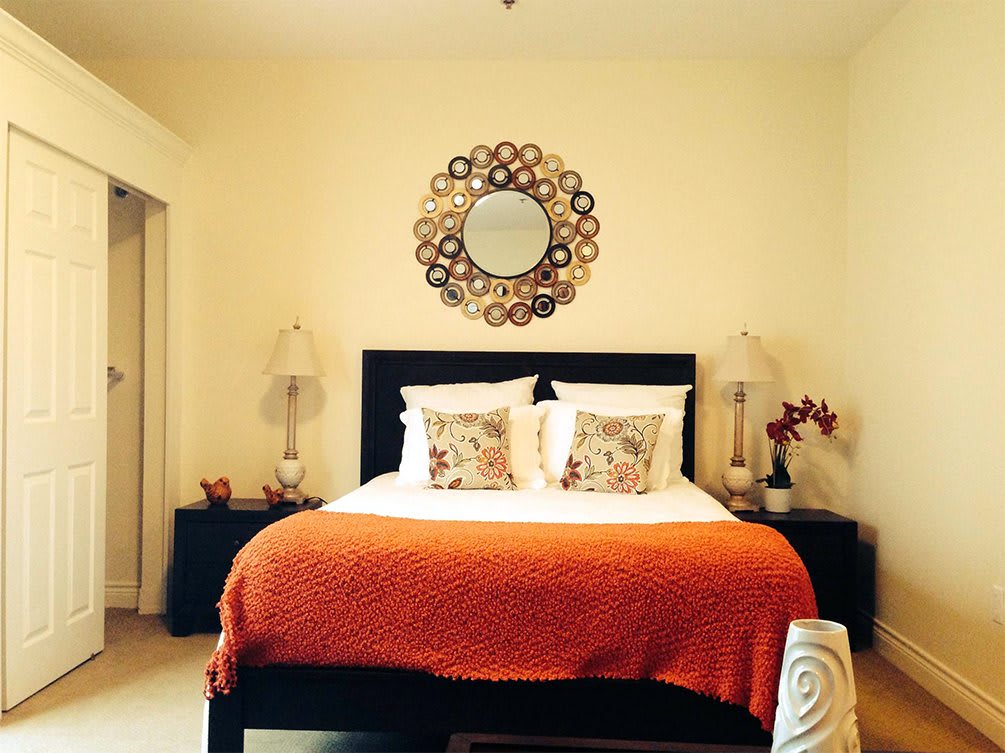A stylish bedroom featuring a central bed with an orange blanket, decorative pillows, and symmetrical lamps. A decorative mirror serves as a focal point above the headboard.