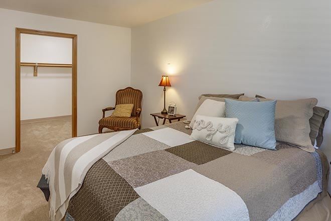 A cozy bedroom with a quilted bedspread, a side table with a lamp, and an upholstered chair.