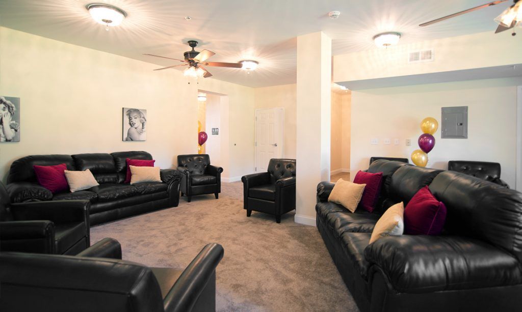 A cozy living room with multiple black leather sofas and colorful throw pillows, featuring a ceiling fan and carpeted floor.