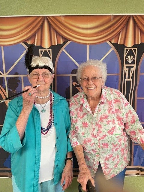 Two elderly women dressed in vintage-themed costumes, smiling for the camera indoors.