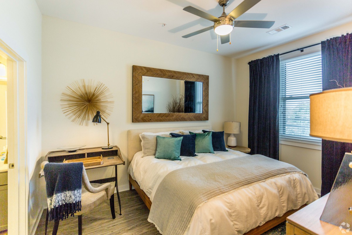A modern bedroom with stylish decor, featuring a bed with multiple pillows, a mirror above the headboard, a desk with a lamp, and a ceiling fan.